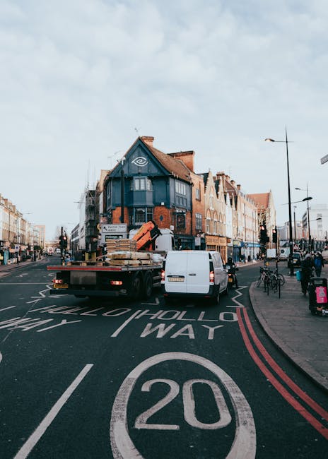 A white flatbed truck loaded with wooden pallets and cardboard boxes is parked on a city street with a 20 mph speed limit marking on the road. The truck is positioned adjacent to a multi-storey building with a blue facade and red brick elements, situated on the corner of a busy area near Lordship Lane and Crown Point in Dulwich. The scene includes pedestrians walking along the sidewalk, bicycles parked on the pavement, and streetlights and traffic signals visible in the background. The parked vehicles and the loading process indicate ongoing house removals or home relocation activities, with Dulwich Removals providing moving services in this residential and commercial area during daylight hours, under a partly cloudy sky.