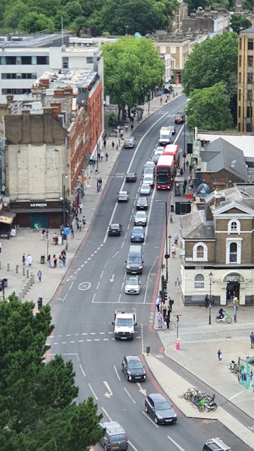 A street scene in Dulwich with parked cars lining both sides of the road, including hatchbacks, sedans, and vans, in front of a historic stone church with a tall, pointed steeple and gothic windows. The street features a mix of residential and commercial buildings, with trees partially obscuring the church and lampposts along the pavement. Two pedestrians, one pushing a bicycle, are walking along the sidewalk, which is strewn with cardboard boxes, packing materials, and a few small furniture items, suggesting a home relocation or moving process. A Dulwich Removals vehicle may be visible in the background near the loading area, highlighting the ongoing furniture transport and packing activities associated with house removals in the area. Bright daylight and a clear sky complete the scene, emphasizing a typical residential move within Dulwich near Lordship Lane and Crown Point, aligning with the page's focus on moving services in SE22, Dulwich.