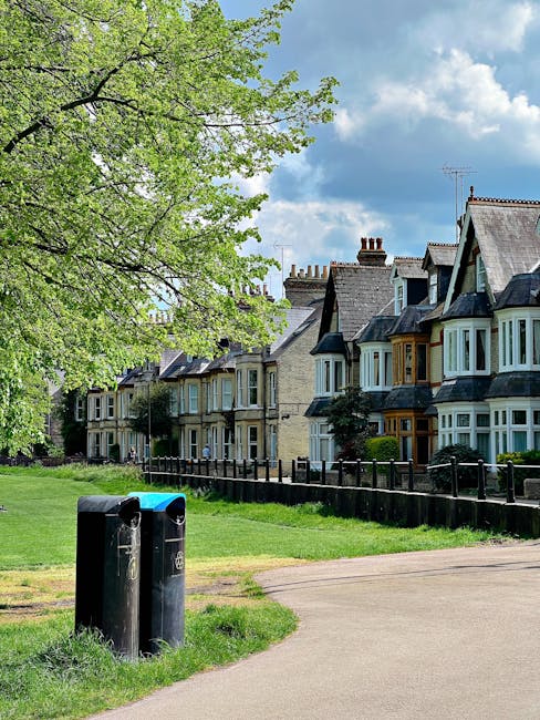 A row of traditional terraced houses with bay windows and slate roofs in Dulwich, visible behind a black metal fence and a well-maintained green lawn. In the foreground, there are two black outdoor trash bins positioned on the pavement near a dirt path. A large leafy tree with bright green foliage extends over the scene from the left side, partially shading the view of the houses. The sky above is partly cloudy with patches of blue. This setting illustrates a residential area suitable for house removals and relocation services, highlighting the environment where Dulwich Removals operates, with a focus on moving logistics such as loading or transporting furniture and belongings across the local estate.