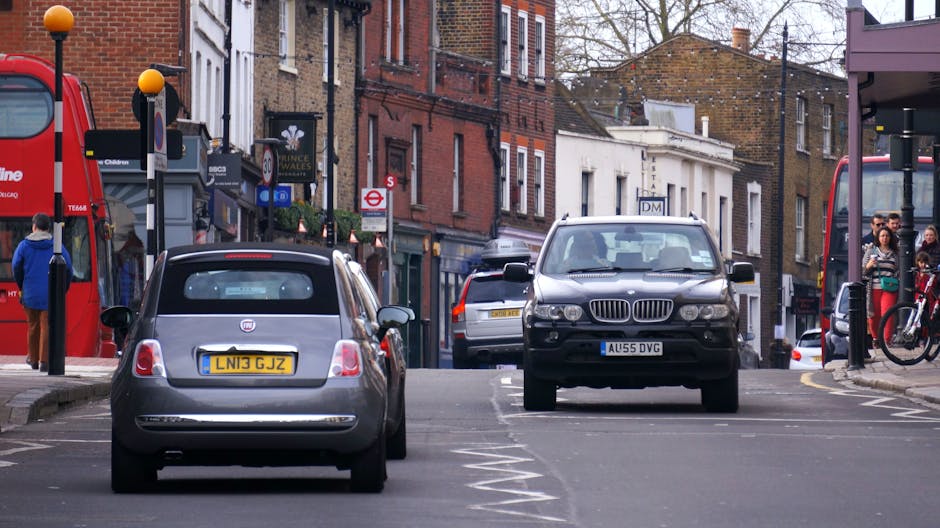 A street scene in East Dulwich showing a parked fiat 500 with UK license plates in the foreground, adjacent to a black BMW SUV that is facing towards the camera. The area appears to be a busy commercial and residential zone with closely spaced brick and stucco buildings lining the street. On the left side, there is a red double-decker bus stopping at a bus stop, and a few pedestrians are waiting nearby, some standing on the pavement and others walking. The pavement has a mixture of concrete slabs and brickwork, with a designated parking lane marked by white dashed lines. Behind the parked vehicles, additional cars and a van are visible, indicating typical urban traffic. Several shopfronts with signage and storefront displays are lined along the street, including a small shop with a dark green awning and a sign. Public transportation signs, street lamps, and traffic signs are also visible, contributing to the urban environment. This scene may be representative of a typical day during a home or furniture removal process, where vehicles are parked temporarily for moving logistics, with Dulwich Removals offering services in such busy city areas.