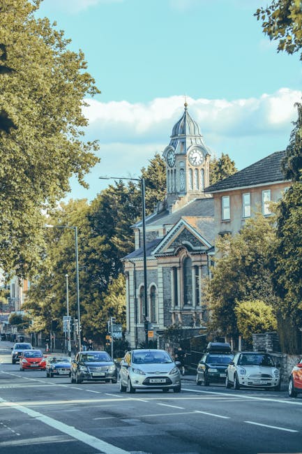 A wide street scene in Dulwich featuring a mix of parked and moving cars along the roadway, with a historic clock tower and church building prominently visible in the background, surrounded by tall, l